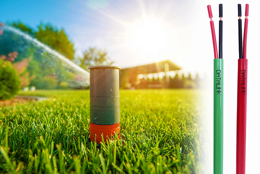 Sistema de irrigação em um jardim verde e exuberante sob a luz do sol brilhante. Um aspersor de irrigação está ativo, esguichando água para o ar, enquanto cabos coloridos vermelho e verde com conectores metálicos estão visíveis no primeiro plano.