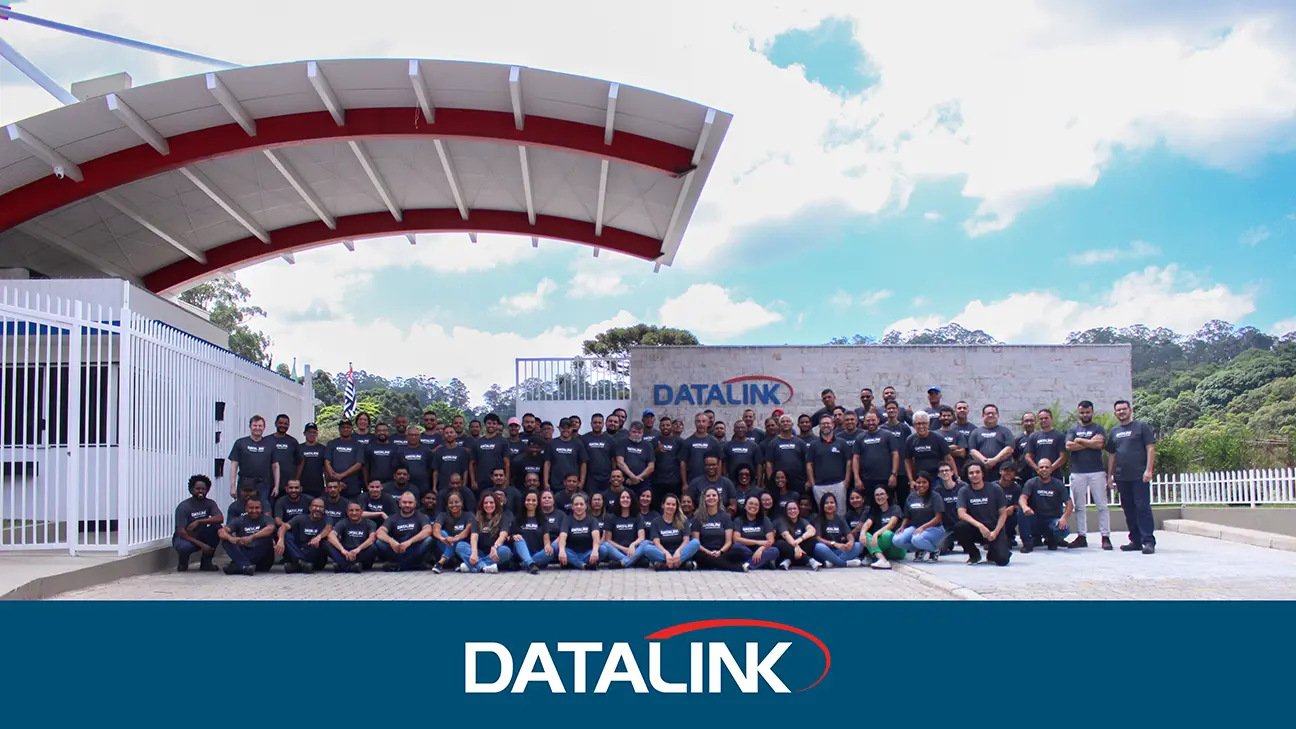 IMG_6815.jpg Fotografia de um grande grupo de pessoas vestindo camisetas escuras, posando em frente à entrada do prédio da Datalink, com uma placa da empresa ao fundo e um céu azul repleto de nuvens acima.