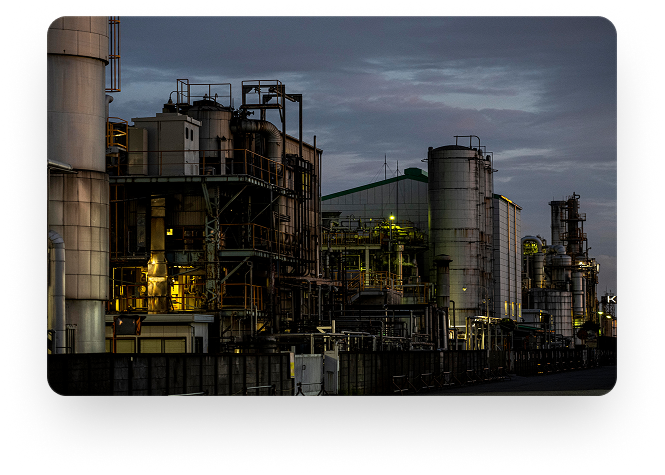 Planta industrial ao entardecer com torres, tanques e tubulações iluminadas.