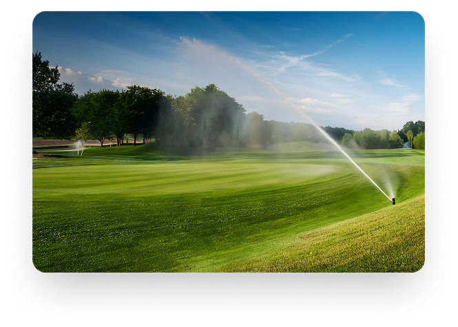 Campo com gramado verde com irrigação ligada com arvores altas ao fundo.