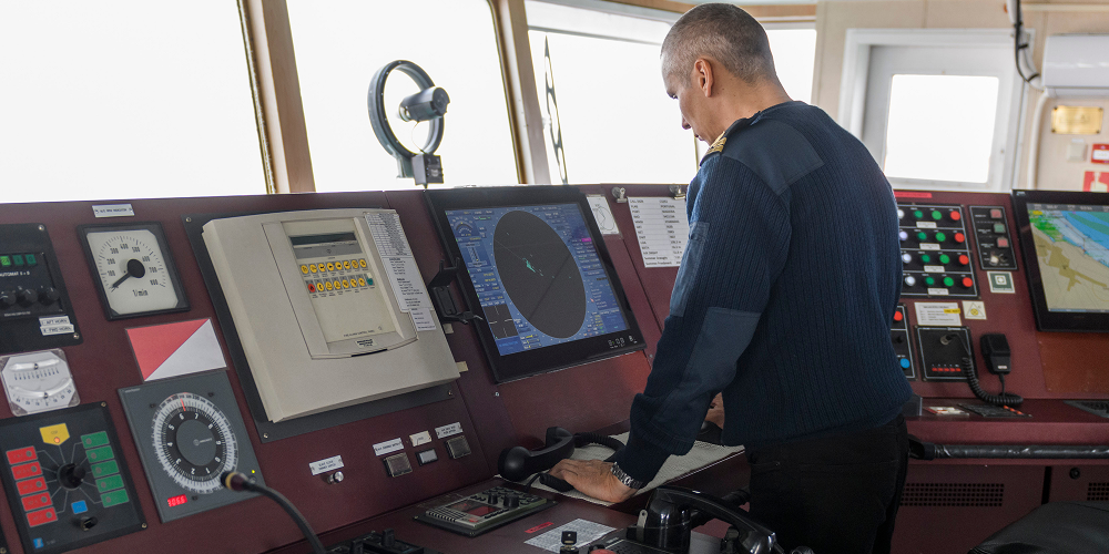 Oficial no passadiço de um navio operando o console de navegação com radar, carta náutica eletrônica e painéis de controle.
