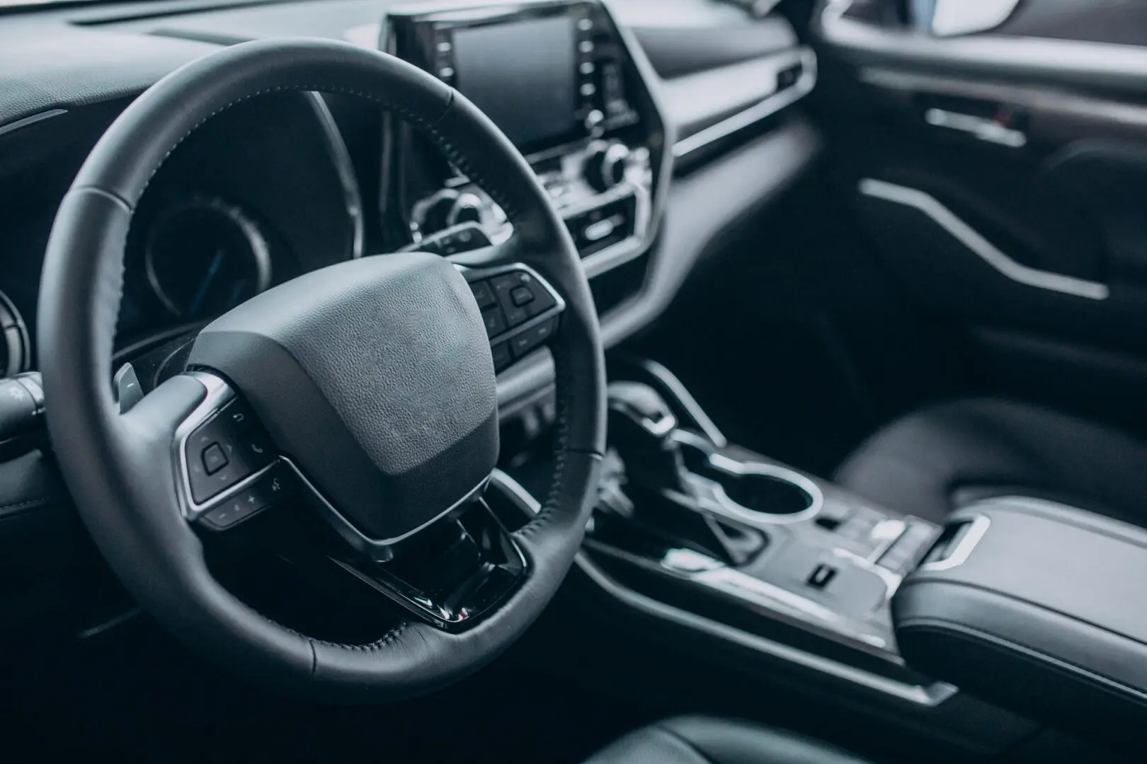 Fotografia mostrando o interior de um carro moderno em tons escuros. Em primeiro plano, vê-se um volante revestido em couro e, ao centro, o painel de controle com tela digital. Mais à direita, ficam a alavanca de câmbio e porta-copos no console central."