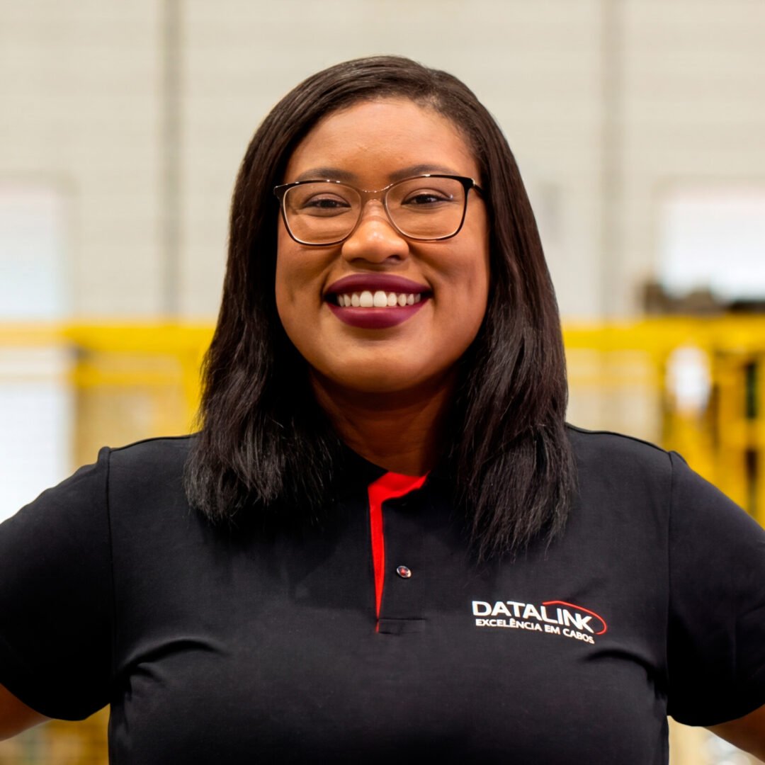 Esta imagem mostra nossa vendendora Jaíne Ferreira vestindo uma camisa polo preta e vermelha com o logotipo “Datalink, Tecnologia em cabos”, em um fundo branco.