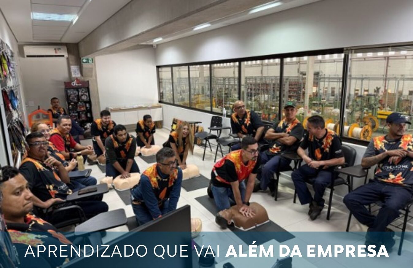 Group of employees in matching vests sit in a training room around CPR mannequins, engaged in a hands-on with an industrial workshop visible through windows in the background.