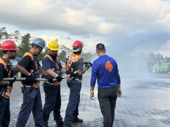 Five trainees in helmets hold hoses while an instructor watches as water sprays on a wet tarmac.